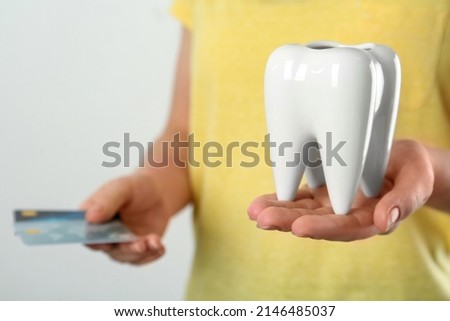 Woman holding ceramic model of tooth and credit cards on light background, closeup. Expensive treatment
