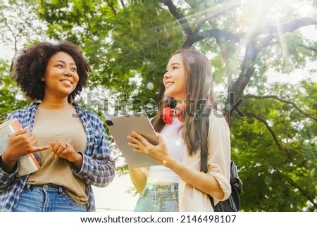 Vivid portrait two ethnic asian and african american female students walking with backpack and school supplies in the nature park watch the beauty tropical plant show their happy faces and eyes. Royalty-Free Stock Photo #2146498107