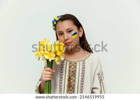 A girl in an embroidered dress stands against a white wall, holding a bouquet of yellow daffodils in her hands.