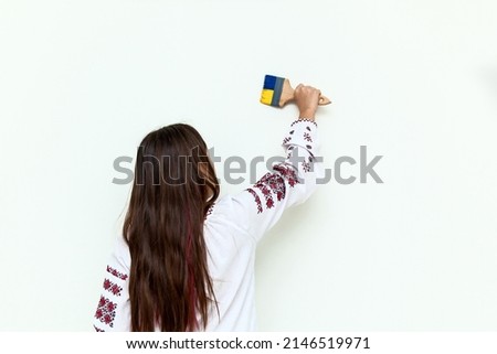 A girl in a dress with embroidery draws on a white wall, holds a brush painted in blue and yellow. The girl protests against the war and draws the flag of Ukraine on the wall