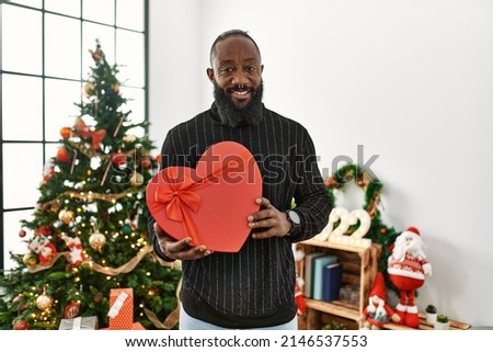 Young african american man holding heart box gift standing by christmas tree at home
