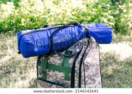 Defocus military backpack and blue tent or sleeping bag. Army bag on green grass background near tree. Military camouflage army rucksack. Tourist summer hiker. Tourism concept. Summer. Out of focus.