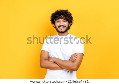 Happy positive indian or arabian curly-haired guy in basic white t-shirt, pointing fingers up at empty mockup space for advertising, announcement, standing on isolated orange background, smiling