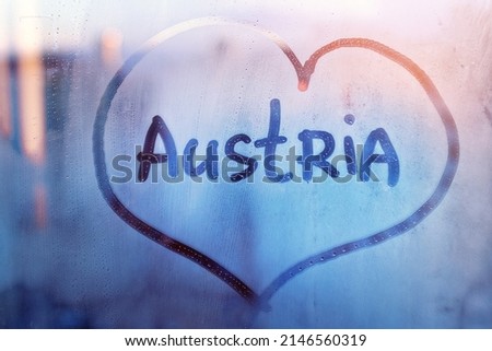 Handwritten word Austria in heart shape on misted blue window with raindrops against background of outlines of urban houses
