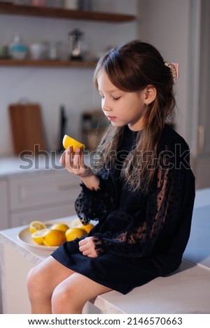Little baby girl trying and showing lemons in kitchen at home. Child in black holiday dress playng and joking with frits. Happiness and joy cooking and living healthy life