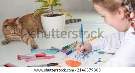 A little girl is drawing, and a cat is lying on the table. High quality photo