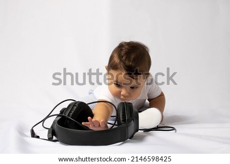 Baby wearing a white shirt and light blue pants with sneakers on a white background with a headset in his hand and expression of curiosity with the unknown object. Babies and technology and music.