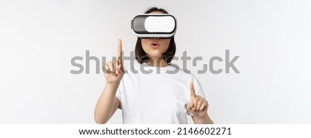 Potrait of asian woman in virtual reality glasses, pointing, choosing smth in VR headset, standing over white background