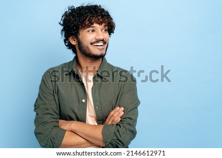 Handsome charismatic successful indian or arabian curly-haired guy in stylish clothes, standing with crossed arms over isolated blue background, looking away, smiling positively Royalty-Free Stock Photo #2146612971