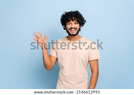 Handsome positive arabian or indian curly-haired guy, wearing casual t-shirt, standing on isolated blue background, looking at the camera, waving hand, welcome sign, smiling friendly