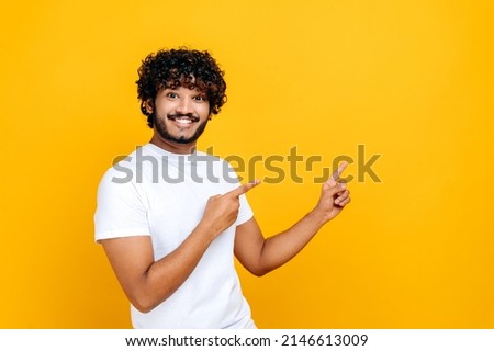 Excited happy indian or arabian guy in a basic white t-shirt, looks at the camera and points fingers to the side, at empty mock-up space, stands on isolated orange background, smiling. Mock-up concept Royalty-Free Stock Photo #2146613009
