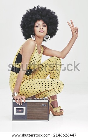 That 70s attitude.... A young woman wearing a 70s retro jumpsuit sitting by a cassette player showing the peace sign. Royalty-Free Stock Photo #2146626447