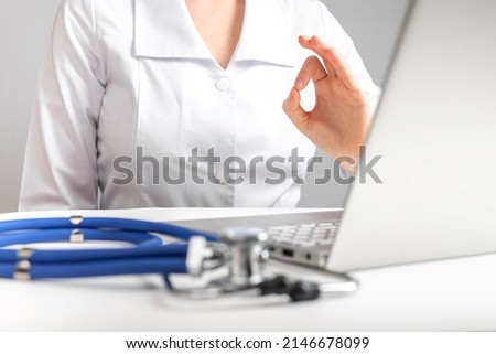 Doctor giving online consultation to patient and showing ok gesture. Telemedicine, telehealth concept. Woman in lab coat sitting at desk with laptop and stethoscope. High quality photo