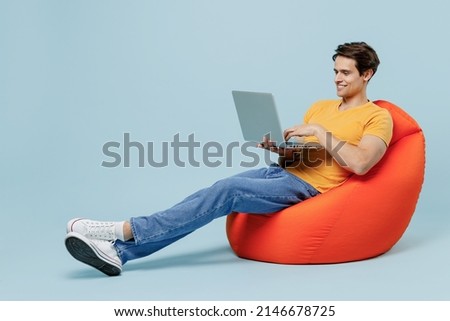 Full body side view fun young man 20s wear yellow t-shirt sit in bag chair hold use work on laptop pc computer isolated on plain pastel light blue background studio portrait. People lifestyle concept