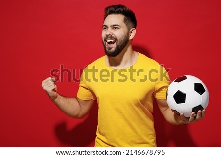 Jubilant fun young bearded man football fan in yellow t-shirt cheer up support favorite team hold soccer ball look aside clenching fists say yes isolated on plain dark red background studio portrait Royalty-Free Stock Photo #2146678795