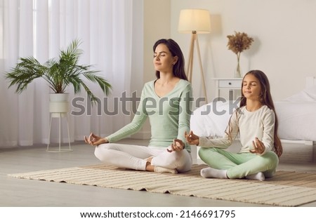 Family yoga. Beautiful young woman and her charming little daughter are smiling while doing yoga together at home. Family sits in lotus position on floor in living room. Mom teaches child to meditate. Royalty-Free Stock Photo #2146691795