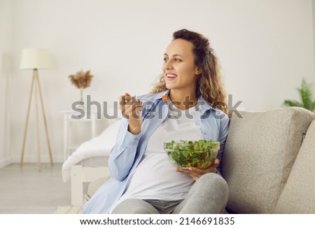 Pregnant woman who eats many different vegetables during pregnancy enjoys fresh green salad at home. Happy woman eating salad for breakfast sitting on sofa in living room. Concept of healthy pregnancy Royalty-Free Stock Photo #2146691835