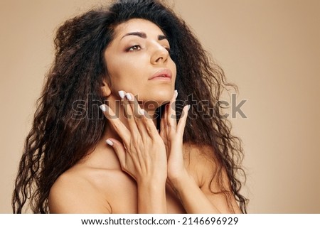 ANTI AGING COSMETIC AD. SKINCARE CONCEPT. Satisfied tanned awesome curly Latin lady deep moisturizing nourishing cream touch cheeks posing isolated on pastel beige background look aside. Cool offer