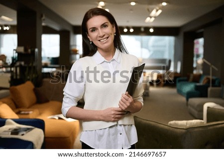 Pleasant European pretty woman, retail assistant and sales rep holding catalog with upholstered furniture, standing in the exposition center and smiling with cheerful toothy smile looking at camera Royalty-Free Stock Photo #2146697657