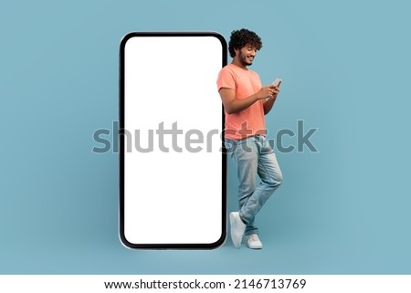 Cool curly indian guy in casual outfit leaning on huge smartphone with white blank screen, using cell phone and smiling, blue studio background, mockup, panorama with copy space