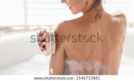 Back view of millennial woman making lymphatic massage with brush, scrubbing skin while taking bubble bath at home, panorama with copy space. Young lady brushing her back in bathtub