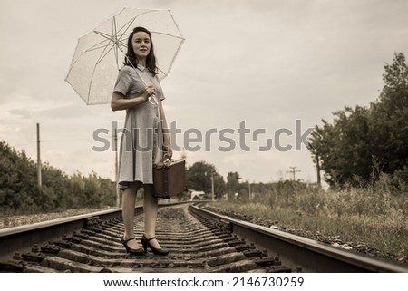 A young fashionista woman in vintage style with a suitcase and a summer umbrella stands on the railway and looks ahead purposefully. Royalty-Free Stock Photo #2146730259