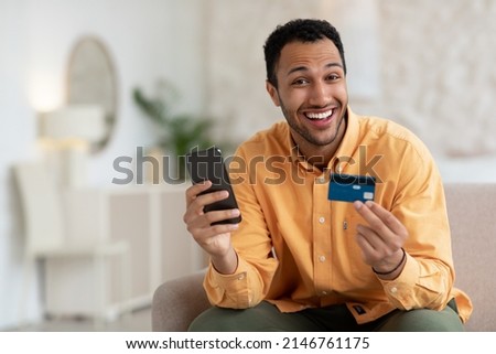 Fast Online Shopping. Smiling young Arab guy holding debit credit card in hand and using cell phone, sitting on the couch at home in living room, posing looking at camera, free copy space mockup