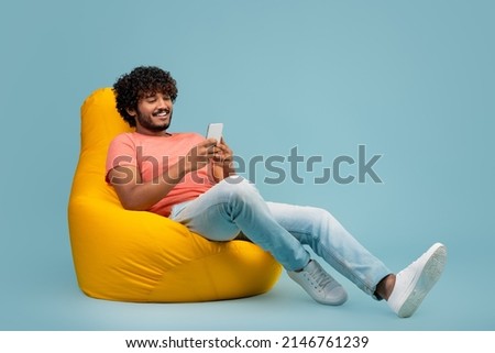Stylish millennial hindu guy with curly hair sitting on yellow bean bag, using modern cell phone and smiling, using entertaining mobile application, blue studio background, panorama with copy space