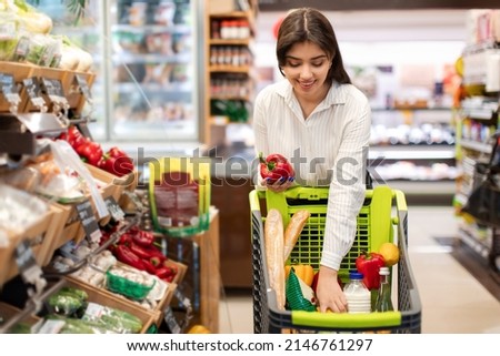 Cheerful Arabic Female Doing Grocery Shopping Putting Red Bell Pepper In Cart Choosing Fresh Vegetables Buying Food In Supermarket On Weekend. Sales And Shop Offers Concept Royalty-Free Stock Photo #2146761297