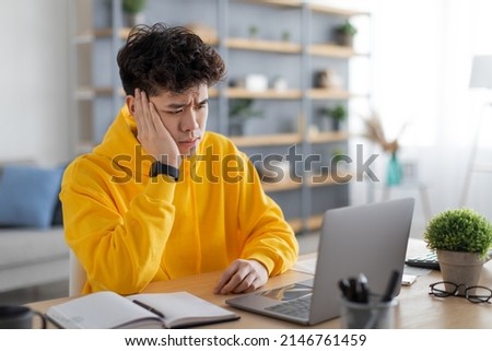 Bored sad young Asian guy sitting at desk using laptop, looking at pc screen, thinking about problems during online lesson or remote work at home office. Tired man leaning head on hand, watching news