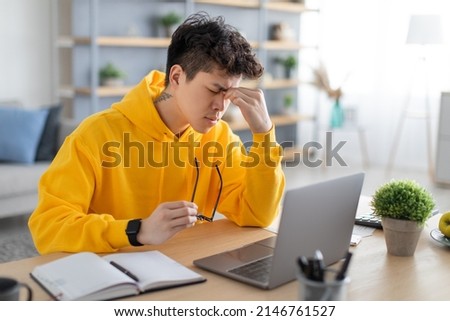 Tired young Asian male freelancer working from home office, using laptop, free copy space. Fatigued guy sitting at desk in front of pc computer, holding taking off glasses, rubbing nose bridge Royalty-Free Stock Photo #2146761527