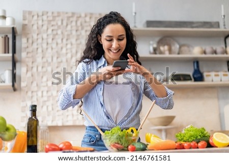 Food blogging concept. Latin woman taking photo of healthy dinner on cellphone, cooking fresh salad while standing in kitchen. Nutrition and weight loss blog concept