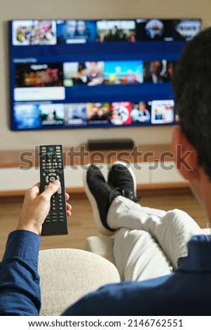 image from behind of unrecognizable man sitting on the sofa with his legs stretched out looking for content to watch on tv Royalty-Free Stock Photo #2146762551
