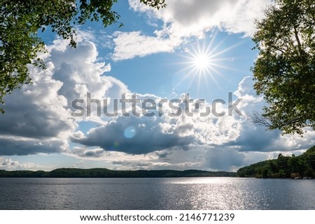 Dramatic sky over forest Ontario Lake in summer. Cumulus clouds surround the sun with showers ahead.  Royalty-Free Stock Photo #2146771239