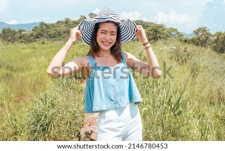 Beautiful cheerful Asian female traveler wearing casual fashionable clothes, hat, standing in outdoor green field or park, smiling with happiness while traveling in summer holiday vacation