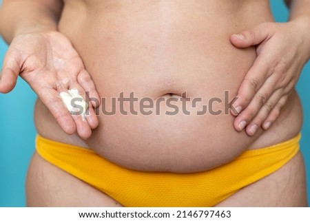 Woman with sagging belly in yellow panties hold body cream on hand closeup. Doing abdominal massage and lifting for weight loss, blue background. Anticellulite fat burning procedure for slim figure.