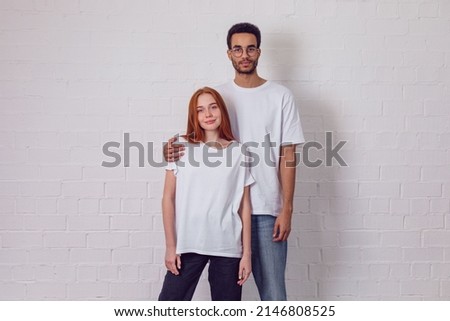 Beautiful international couple in white t-shirts stand on a white background. Mock-up.