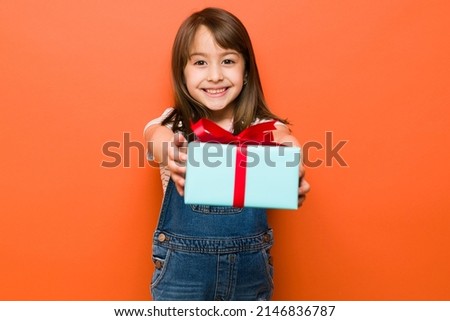 Cheerful brunette girl handing over a gift box with a big smile in a studio against an orange background