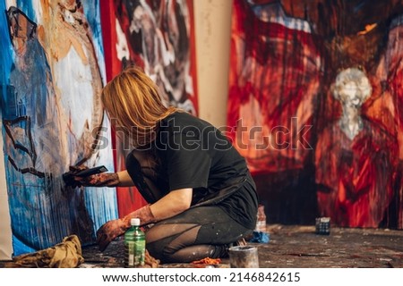 Concentrated woman artist with red hair creating big canvas painting while having a creative process in her studio. Smiling painter being busy with her work at workshop. Art, creativeness concept.