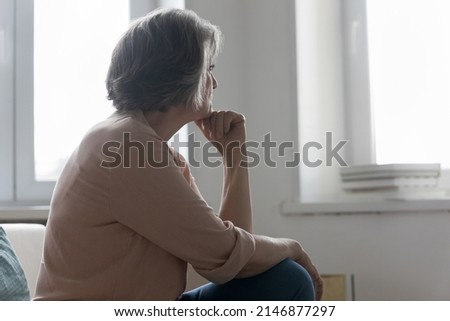 Serious older 50s woman looks out window spend time alone at home, feels sad or disappointed, waits or misses someone seated on sofa indoor. Loneliness, solitude on retirement, nostalgic mood concept Royalty-Free Stock Photo #2146877297