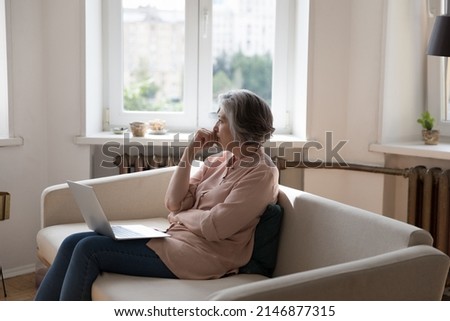 Pensive older woman sits on couch with laptop thinks, staring out window, looks thoughtful or concerned distracted from tech usage, experiences difficulties with modern device, search solution concept Royalty-Free Stock Photo #2146877315