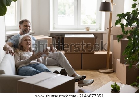 Mature couple using digital tablet buying goods for new home relaxing on cozy couch in living room with packed cardboard boxes with stuff on relocation day. Do easy comfort remote e-shopping concept Royalty-Free Stock Photo #2146877351