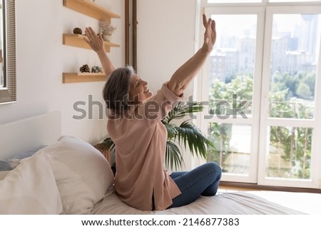 Side view older woman sit on bed in modern light warm bedroom raise hands stretch body muscles feels happy, welcomes new day, enjoy carefree morning, untroubled retired life and comfort living concept Royalty-Free Stock Photo #2146877383