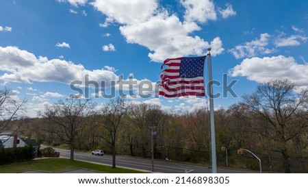 An aerial view of the American flag on a beautiful, sunny day with blue skies and white clouds. The flag blows in the wind, just above the tree line of a park on Long Island, New York.