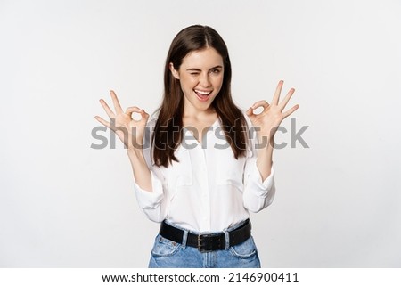 Smiling confident business woman, winking, showing okay ok sign, excellent, no problem gesture, standing over white background