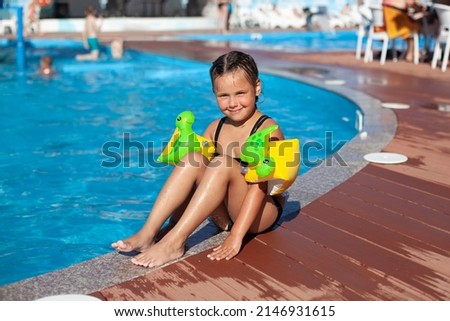 Child poses by pool. Charming happy girl in inflatable swimming armbands with picture of dinosaurs sits on granite side of bassen in water park and looks at camera. 