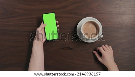 Top view man drinking hot chocolate and using smartphone with green screen over black walnut wood table, wide photo