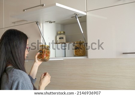 Young Asian woman stacks glass jars with horns in kitchen cabinet. Storage of bulk products. Zero waste. Vegetarian woman. Conscious consumption. Royalty-Free Stock Photo #2146936907