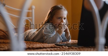 Child watching cartoons on laptop. Serious kid with technology at home on bed spending time on-line. Education and entertainment for toddlers. Girl in cozy atmospheric room over the window