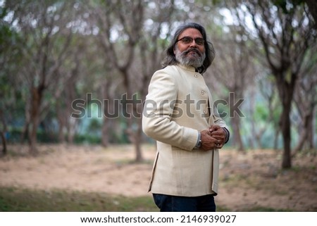 A grey bearded handsome man in band gala traditional handmade woolen coat posing for picture in a public park. 